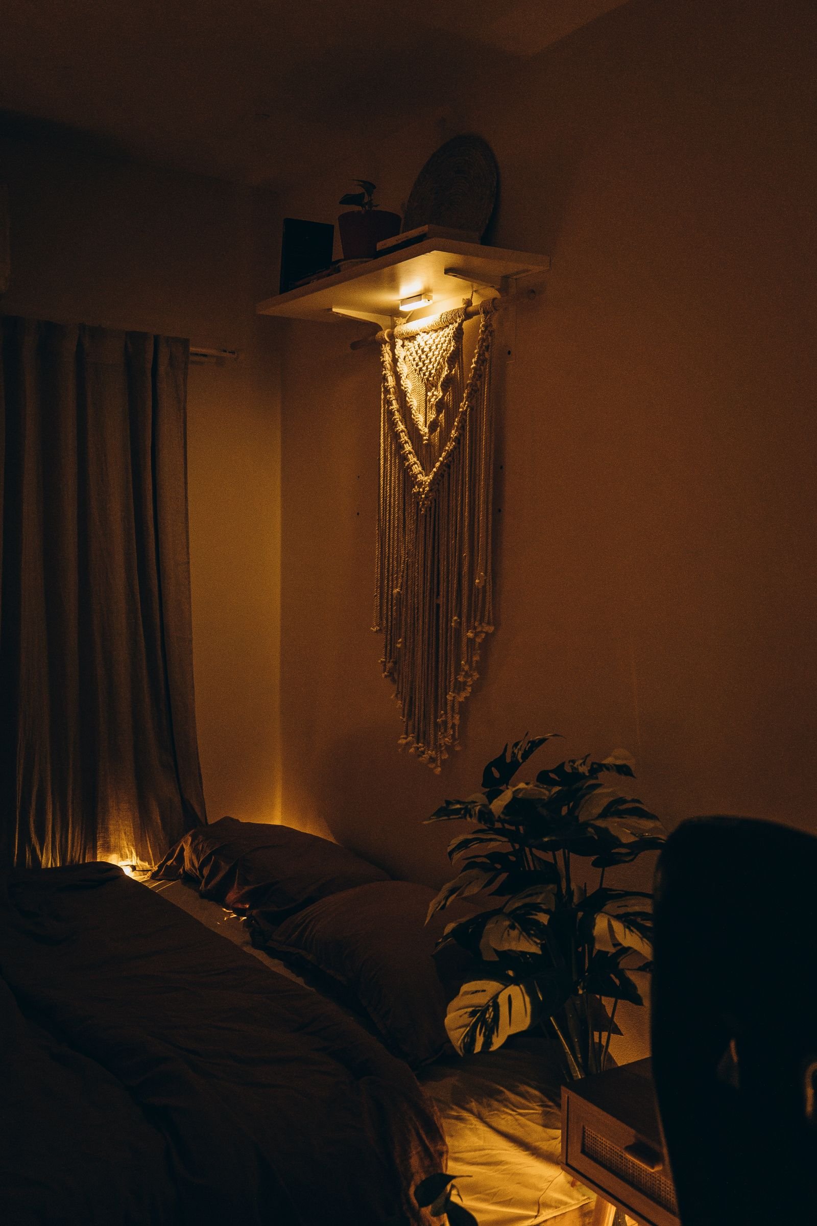 A warm, softly lit bedroom with a macrame wall hanging and a leafy plant beside the bed