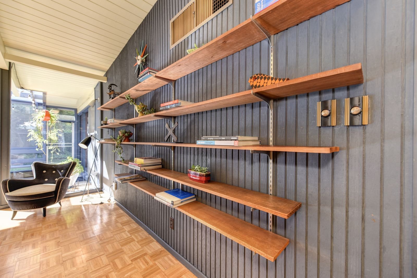 A sunlit living room with wooden floating bookshelves, books, plants and an armchair