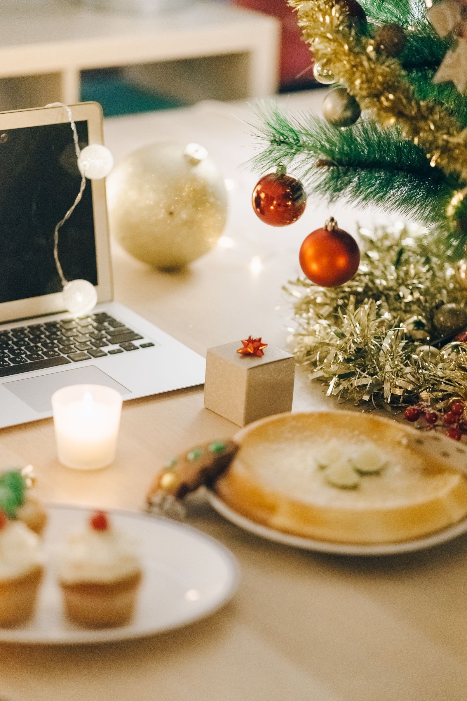 A laptop on a wooden desk beside a lit candle, string lights, red and gold Christmas ornaments and garland