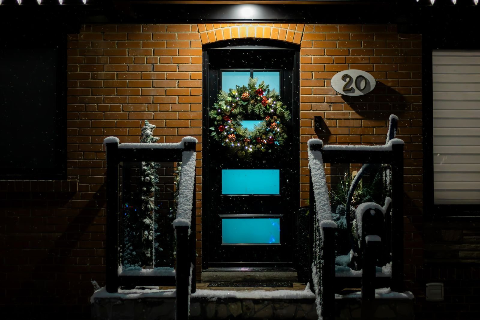 A front door with a lit Christmas wreath and snow-covered steps and railings at night