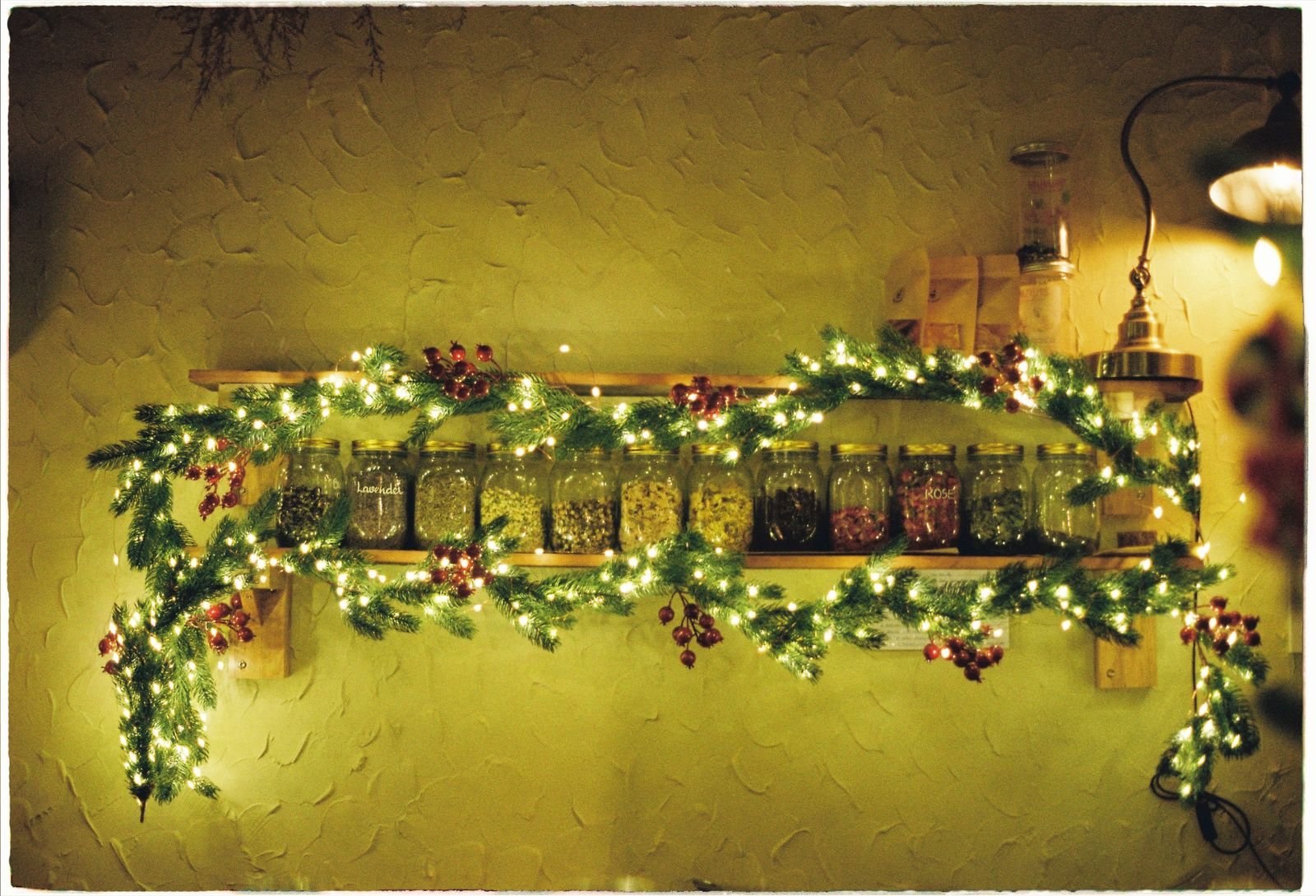 A kitchen shelf decorated with Christmas garland, red berries and warm fairy lights above spice jars