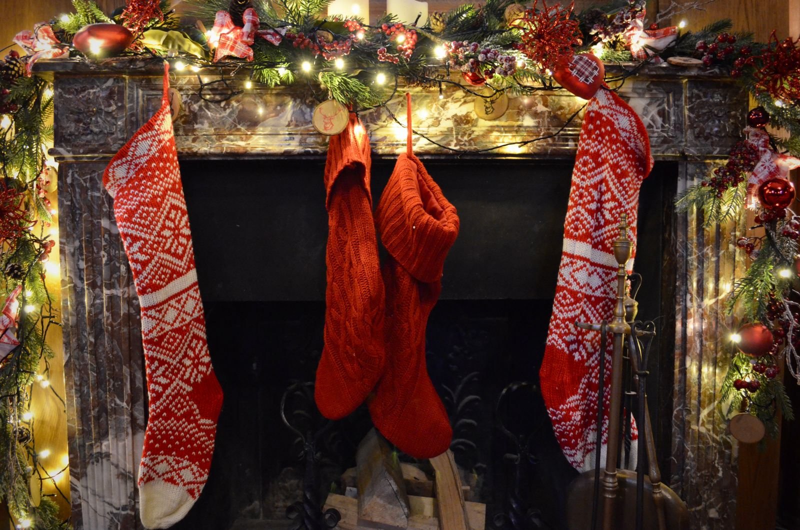 A fireplace mantel with evergreen garland, twinkle lights and red knit Christmas stockings
