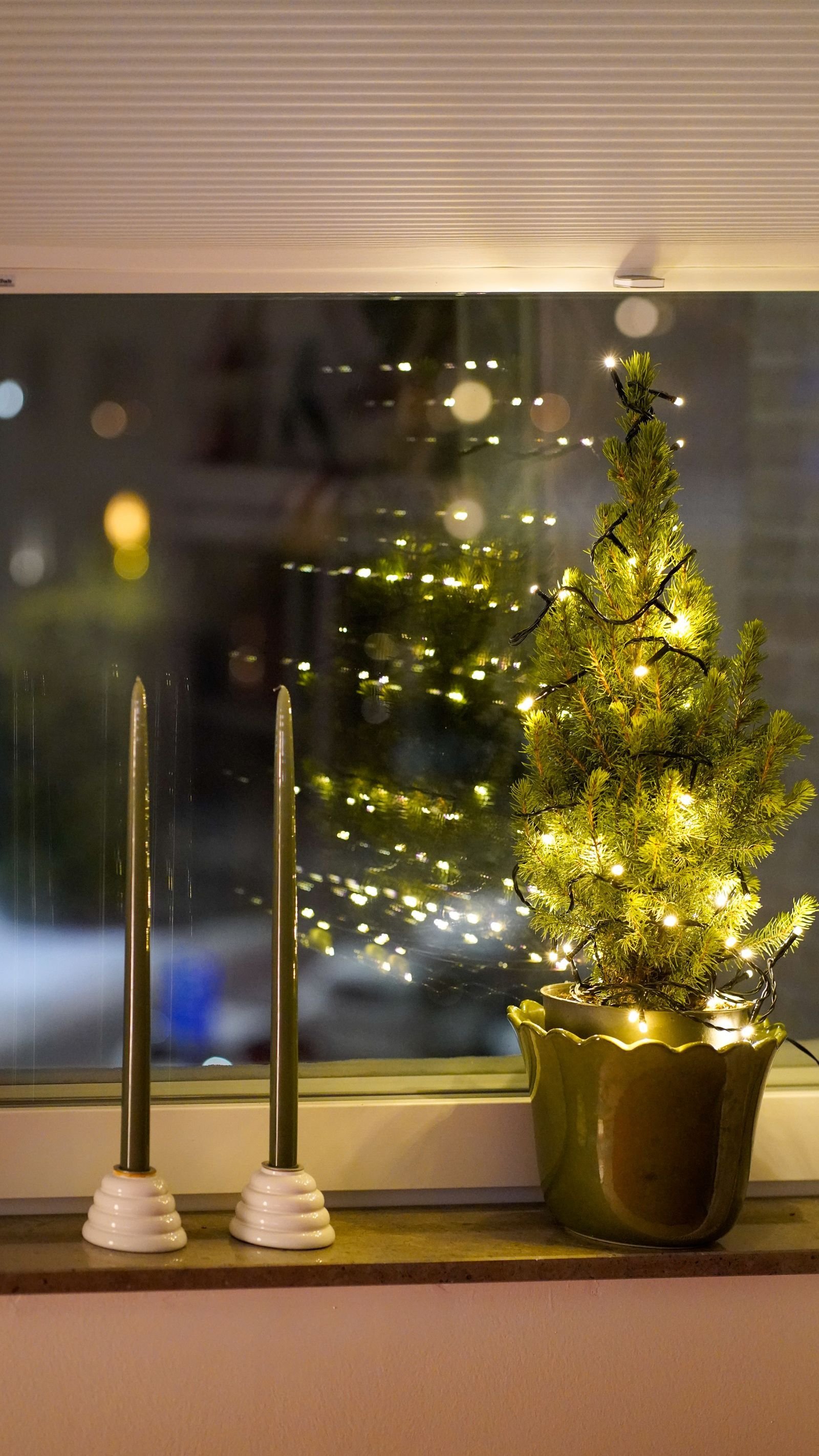 A small lit Christmas tree in a pot and two tall candles on a window ledge at night