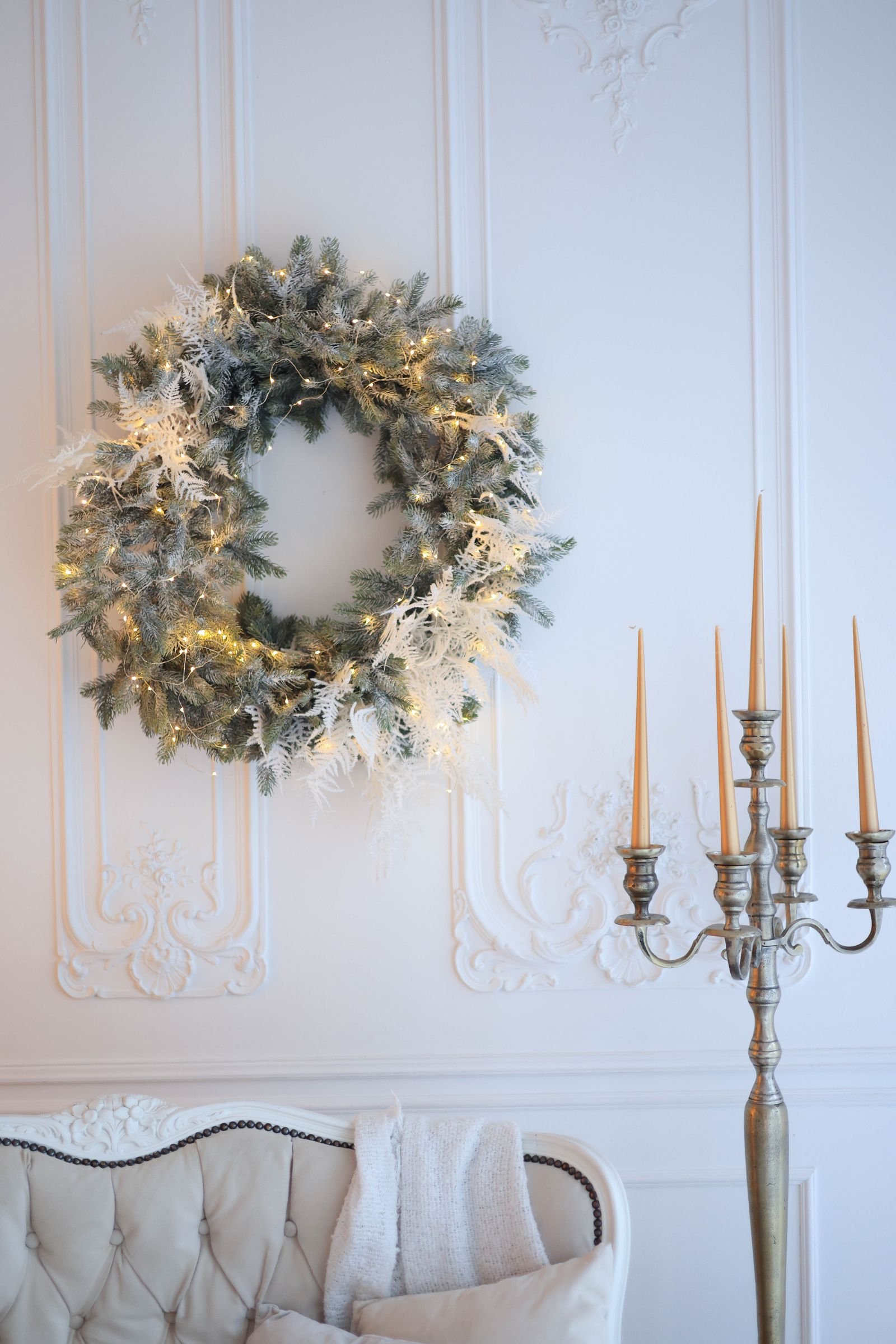 A frosted Christmas wreath with warm lights hanging on a white paneled wall above a candelabra