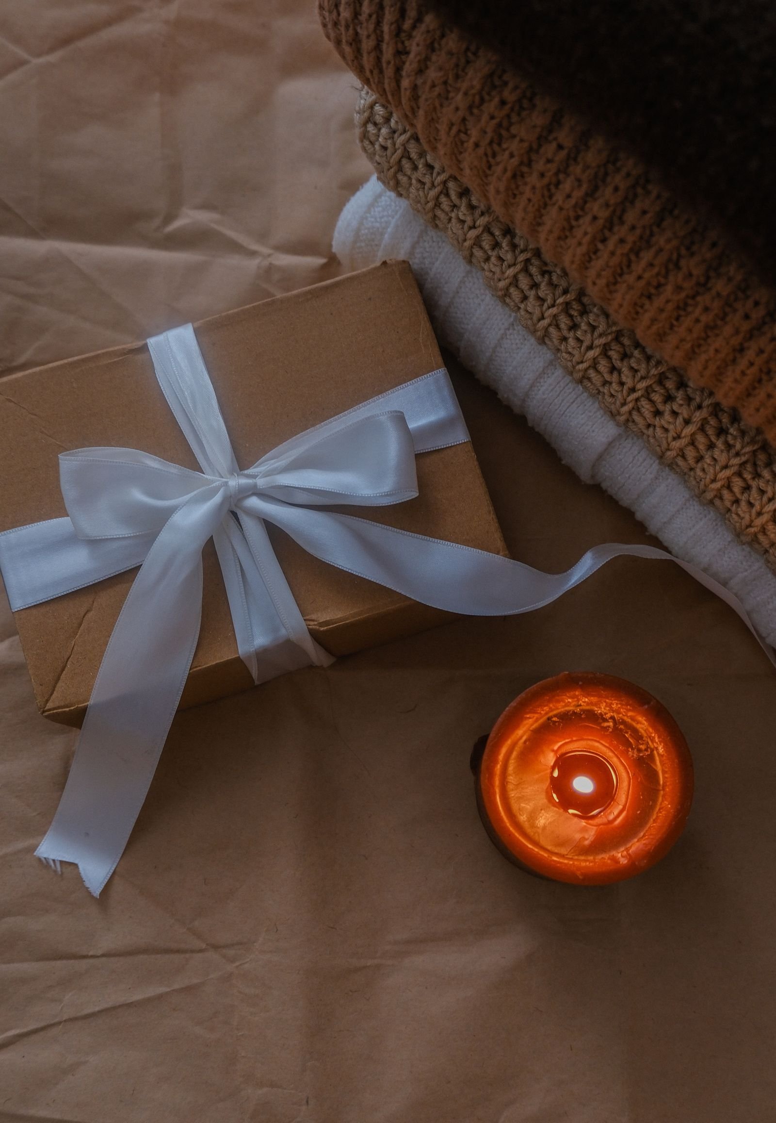 A kraft gift box tied with a pale blue ribbon beside a lit candle and folded knits