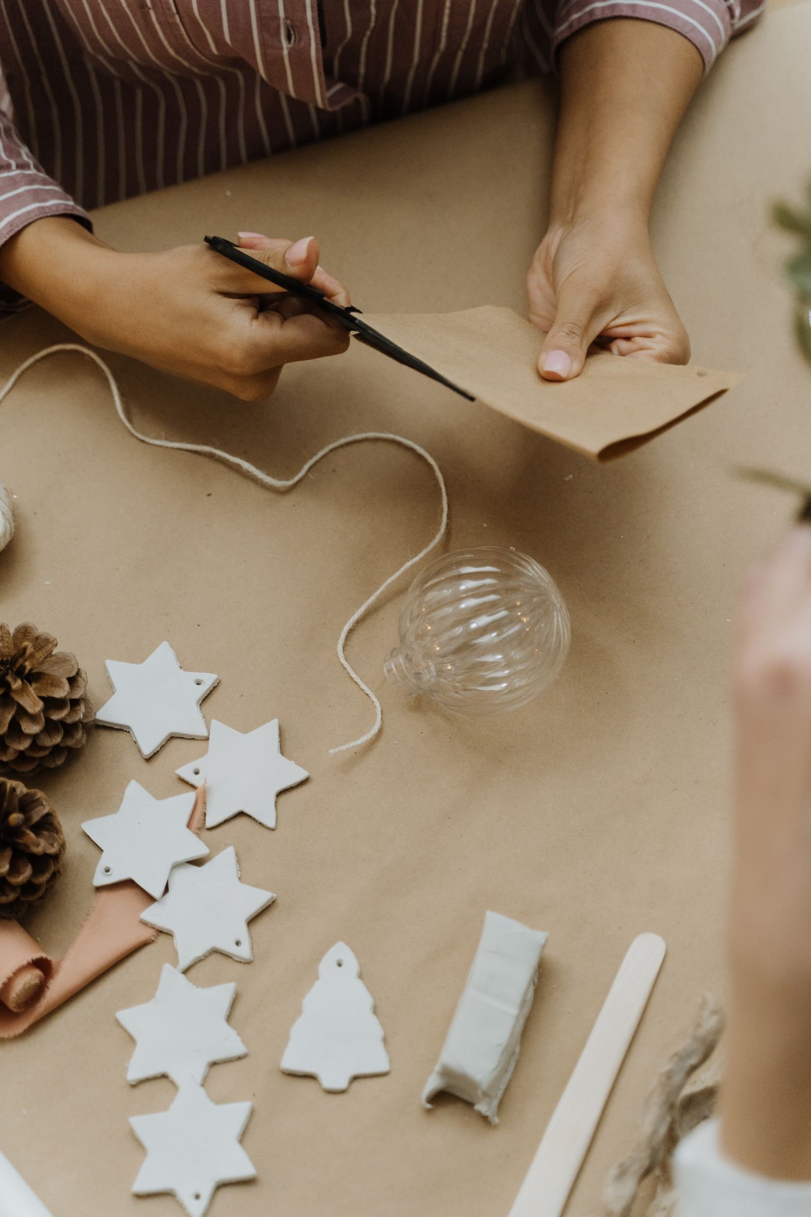 Hands cutting kraft paper next to white clay star ornaments and pinecones for DIY Christmas decor