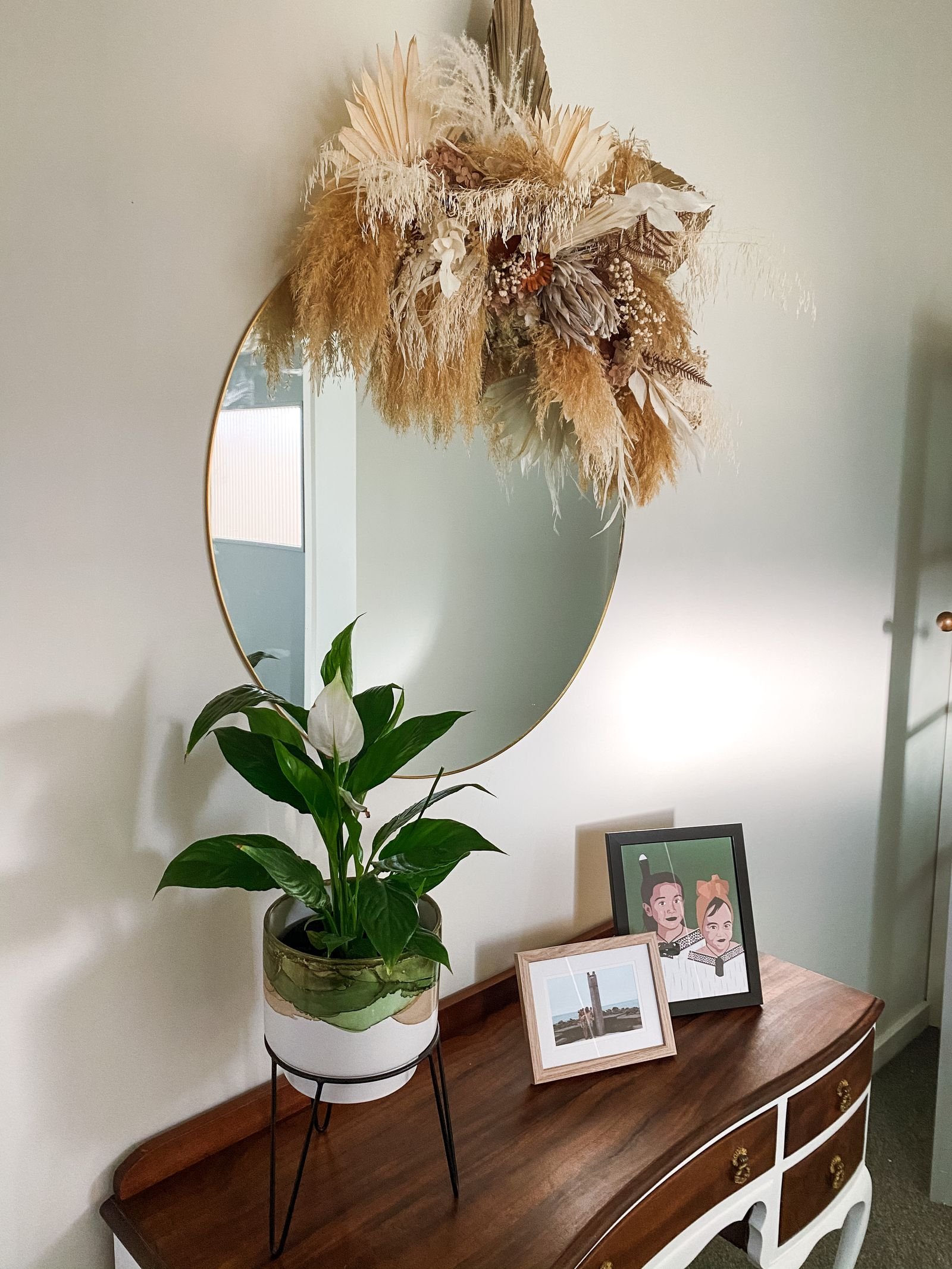 An entryway with a round mirror, a dried pampas arrangement, a plant and framed photos on a wooden console