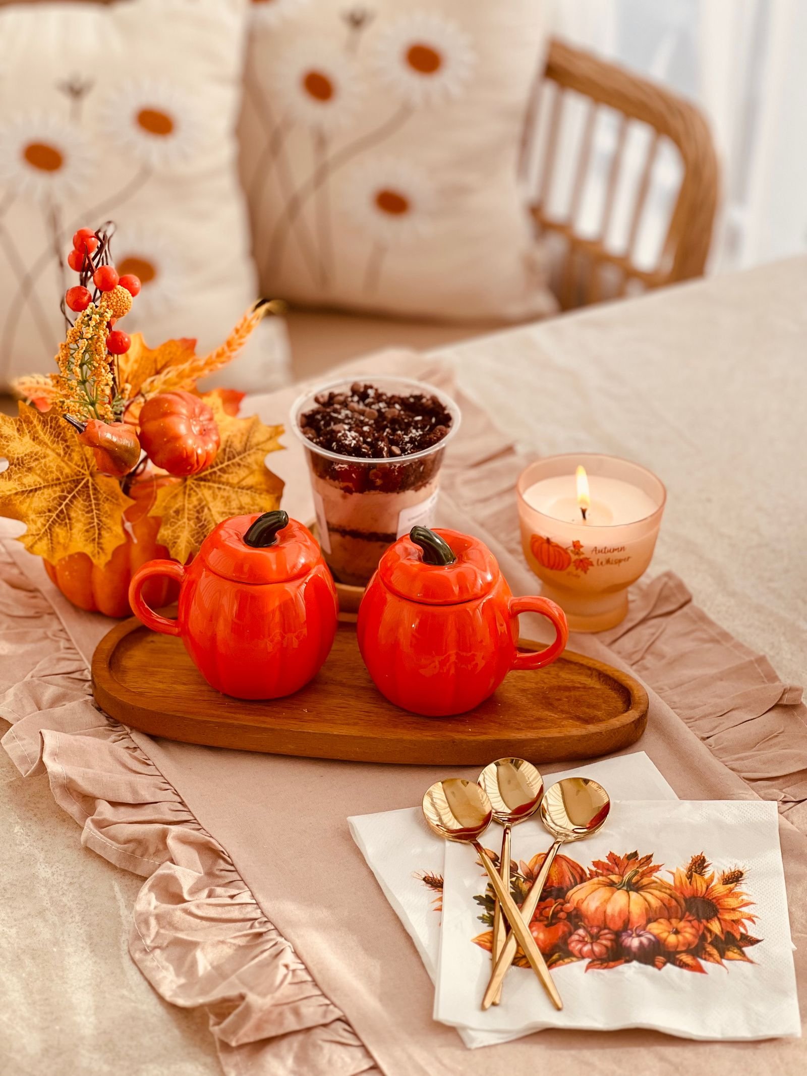 A table set with pumpkin-shaped mugs on a wooden tray, a lit candle, autumn leaves and patterned napkins