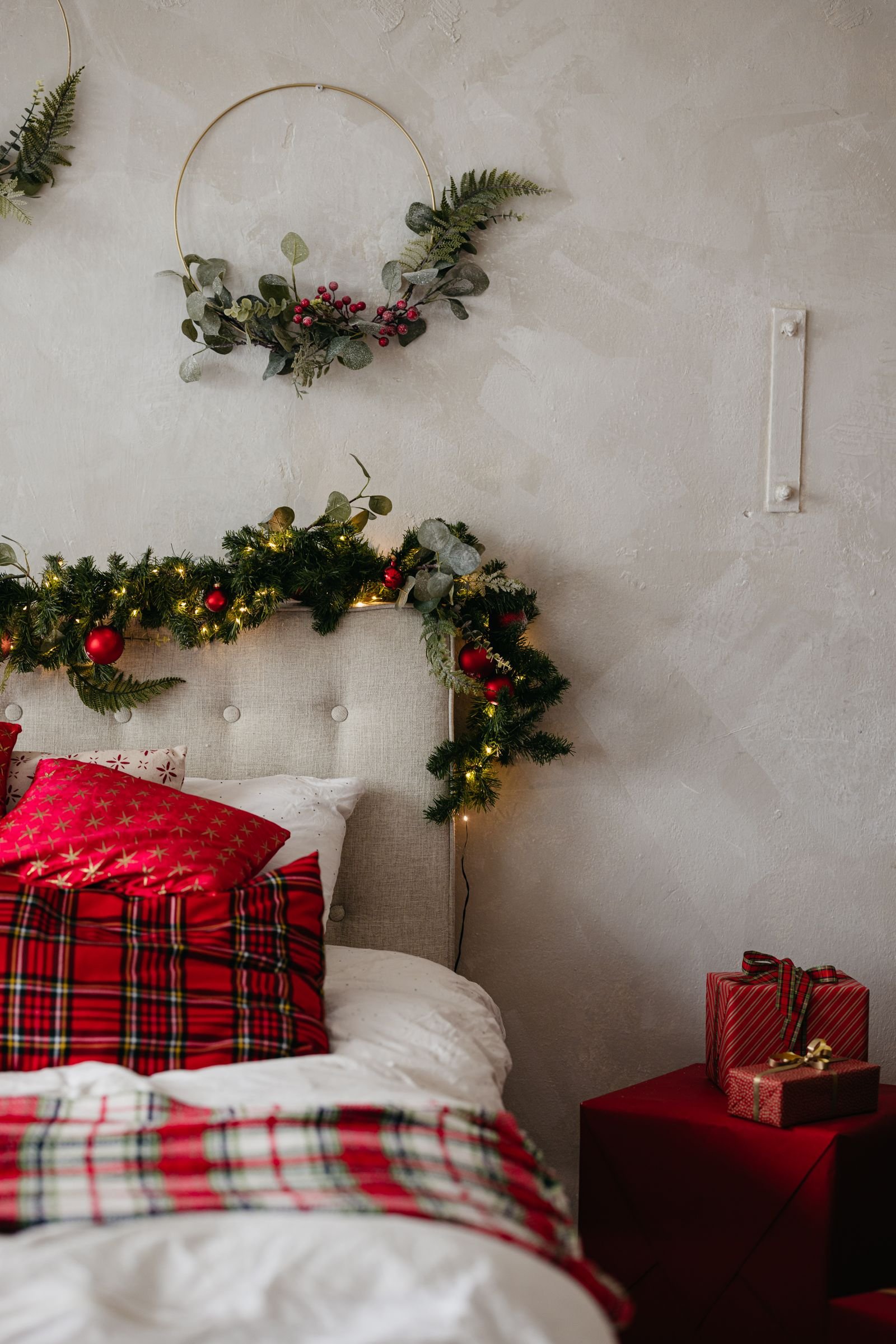 A holiday bedroom with red plaid pillows and blanket, garland on the headboard and a greenery hoop wreath