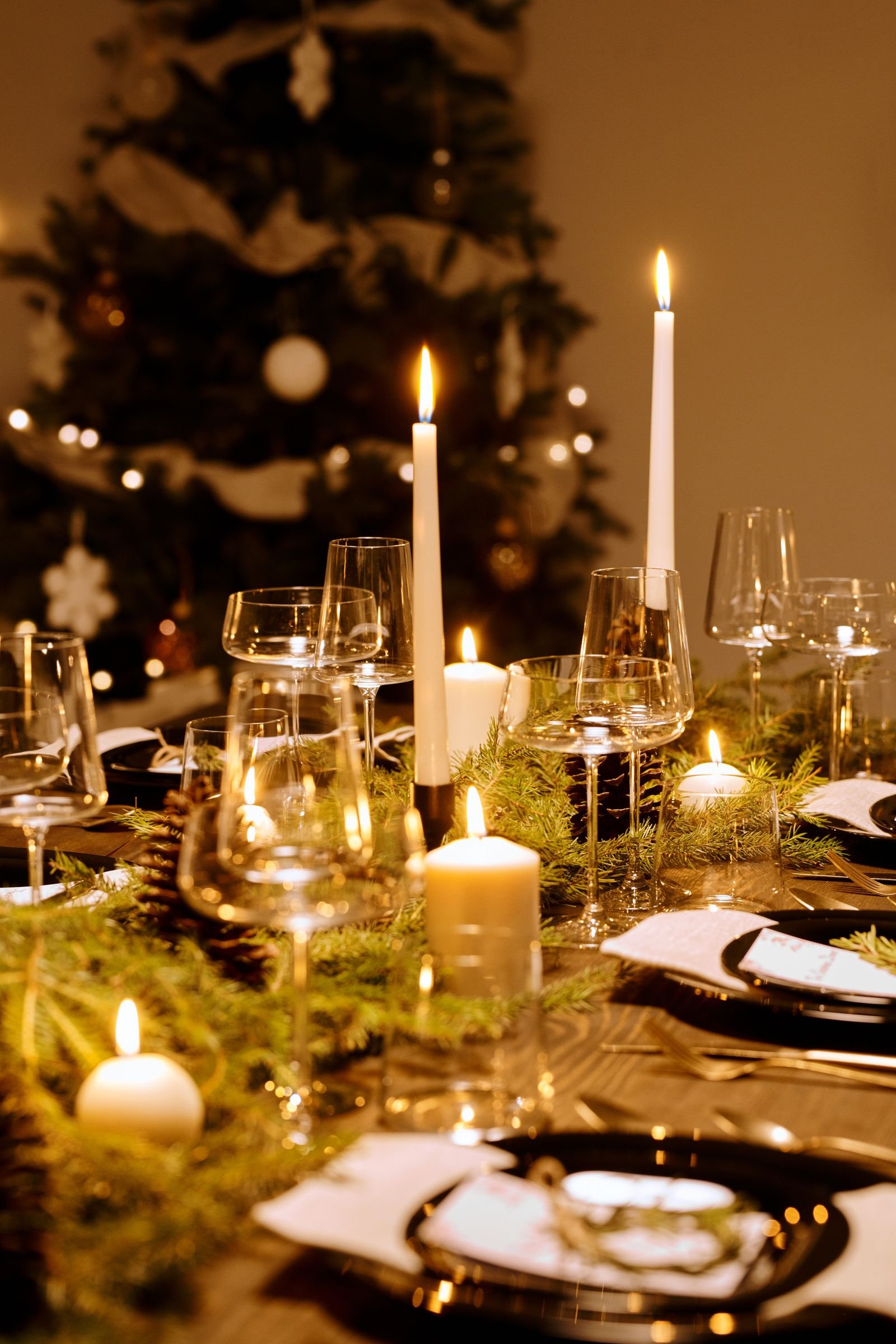 A candlelit holiday dinner table with evergreen sprigs, glassware and a Christmas tree behind