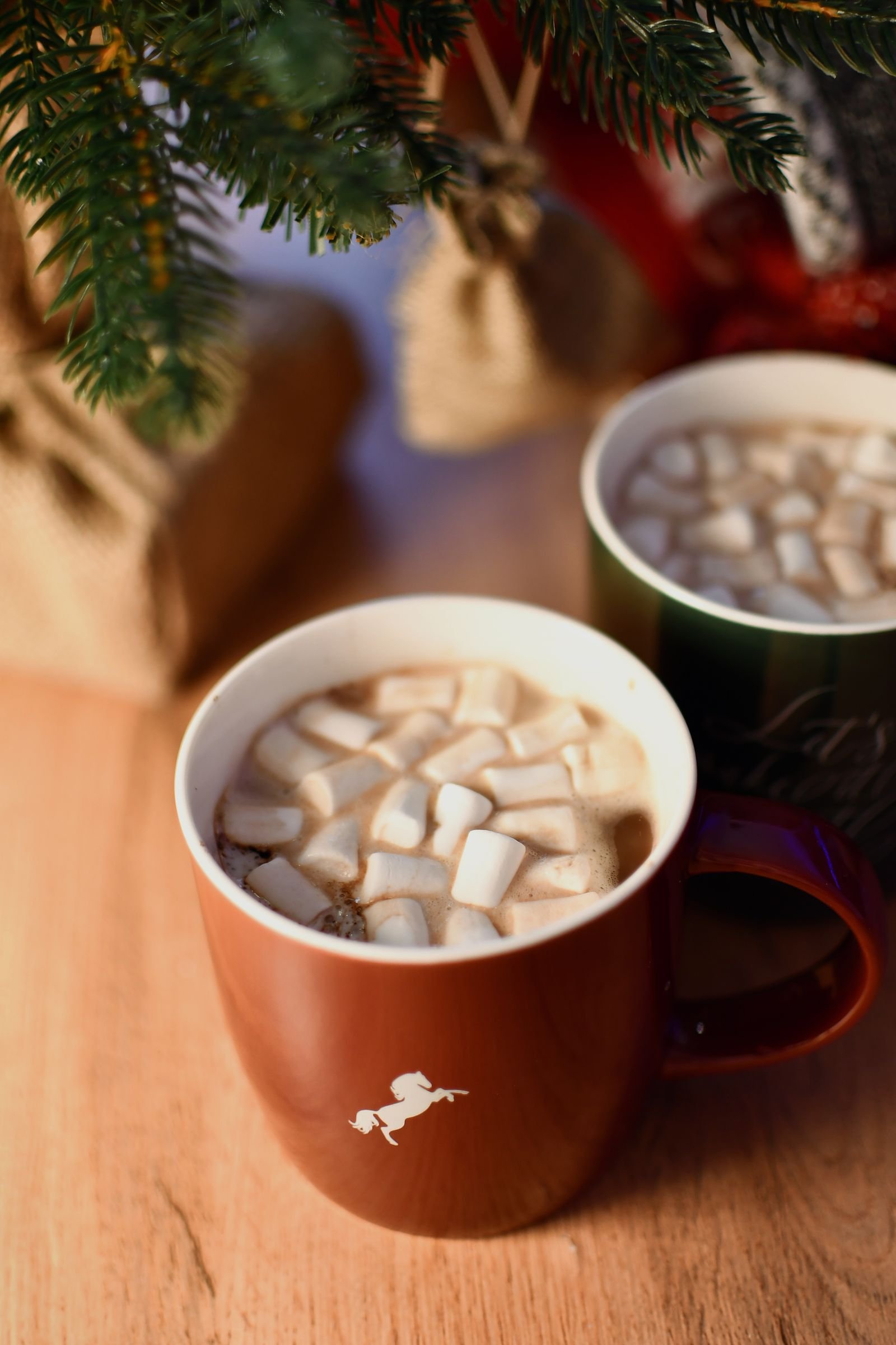 Two mugs of hot chocolate topped with marshmallows on a wooden table beneath Christmas tree branches