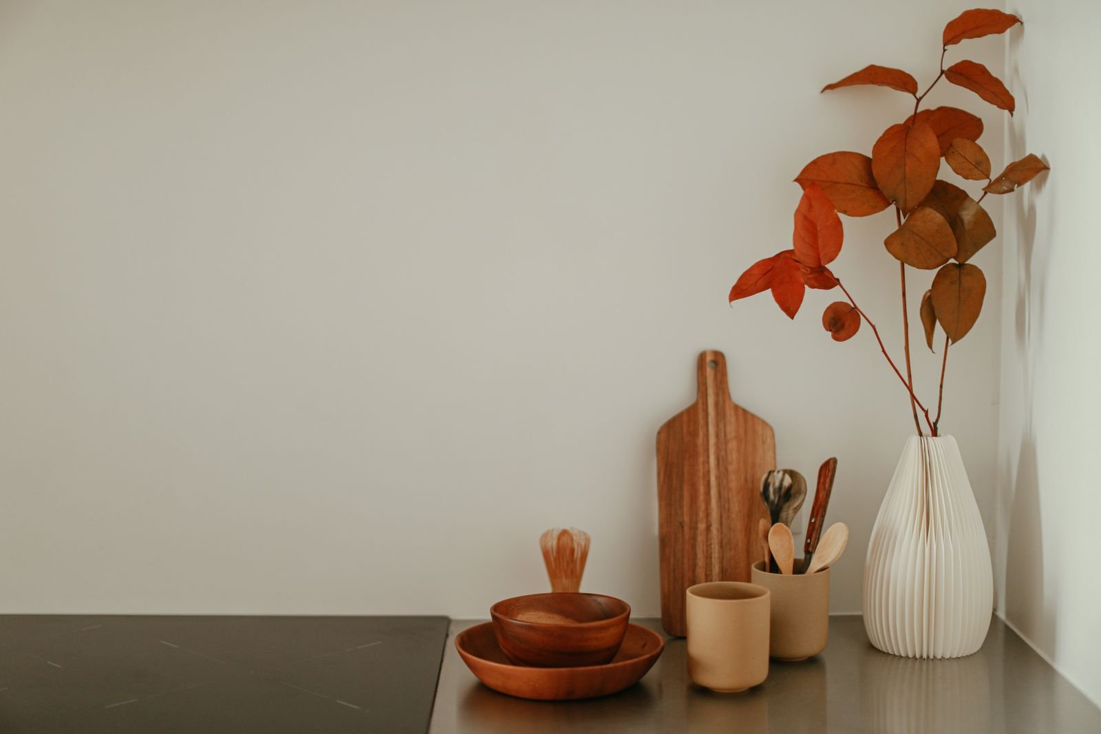 A minimal kitchen counter styled with a wooden cutting board, wooden bowls and a white vase of autumn leaves
