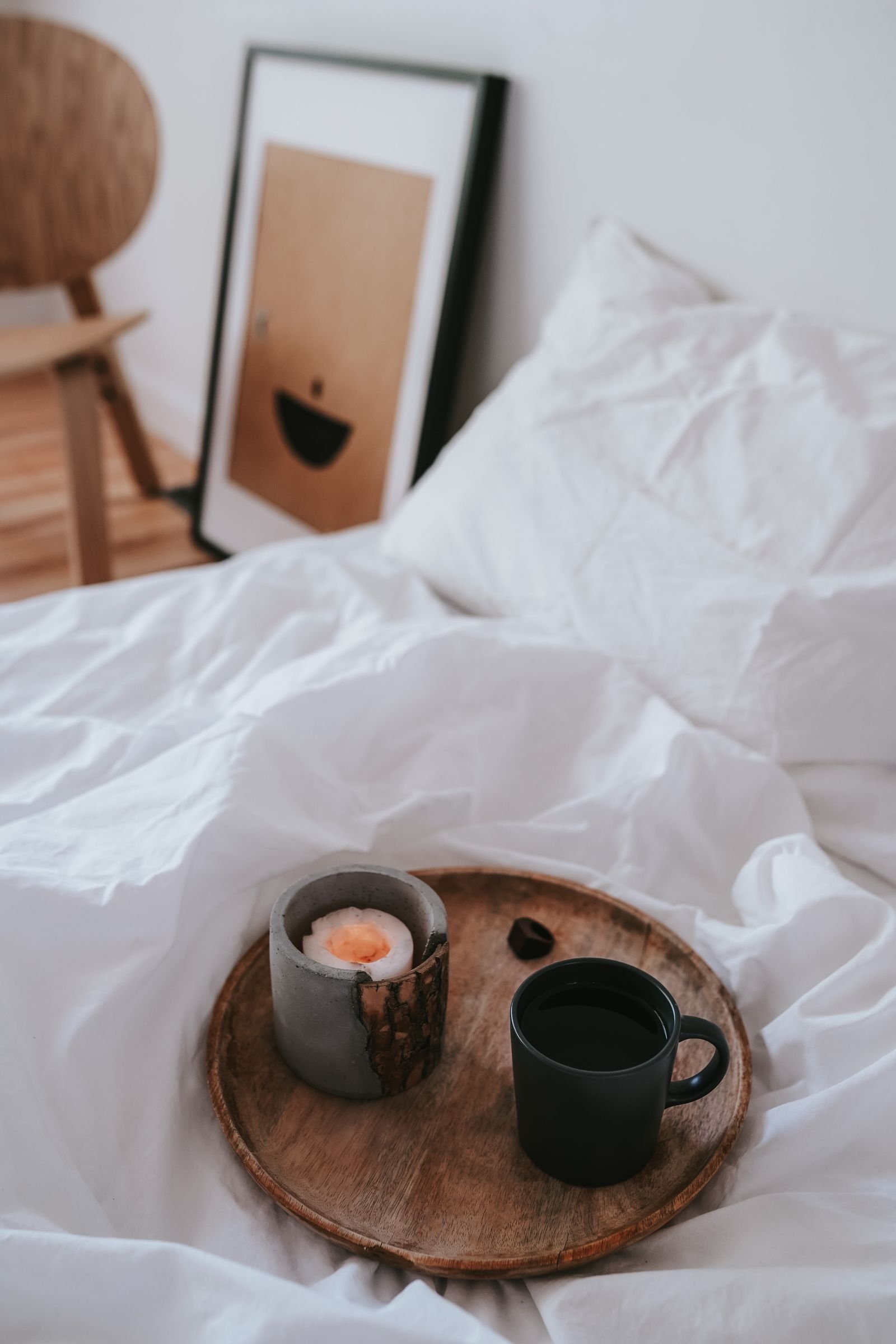 A candle and a ceramic mug on a round wooden tray on a white bed blanket