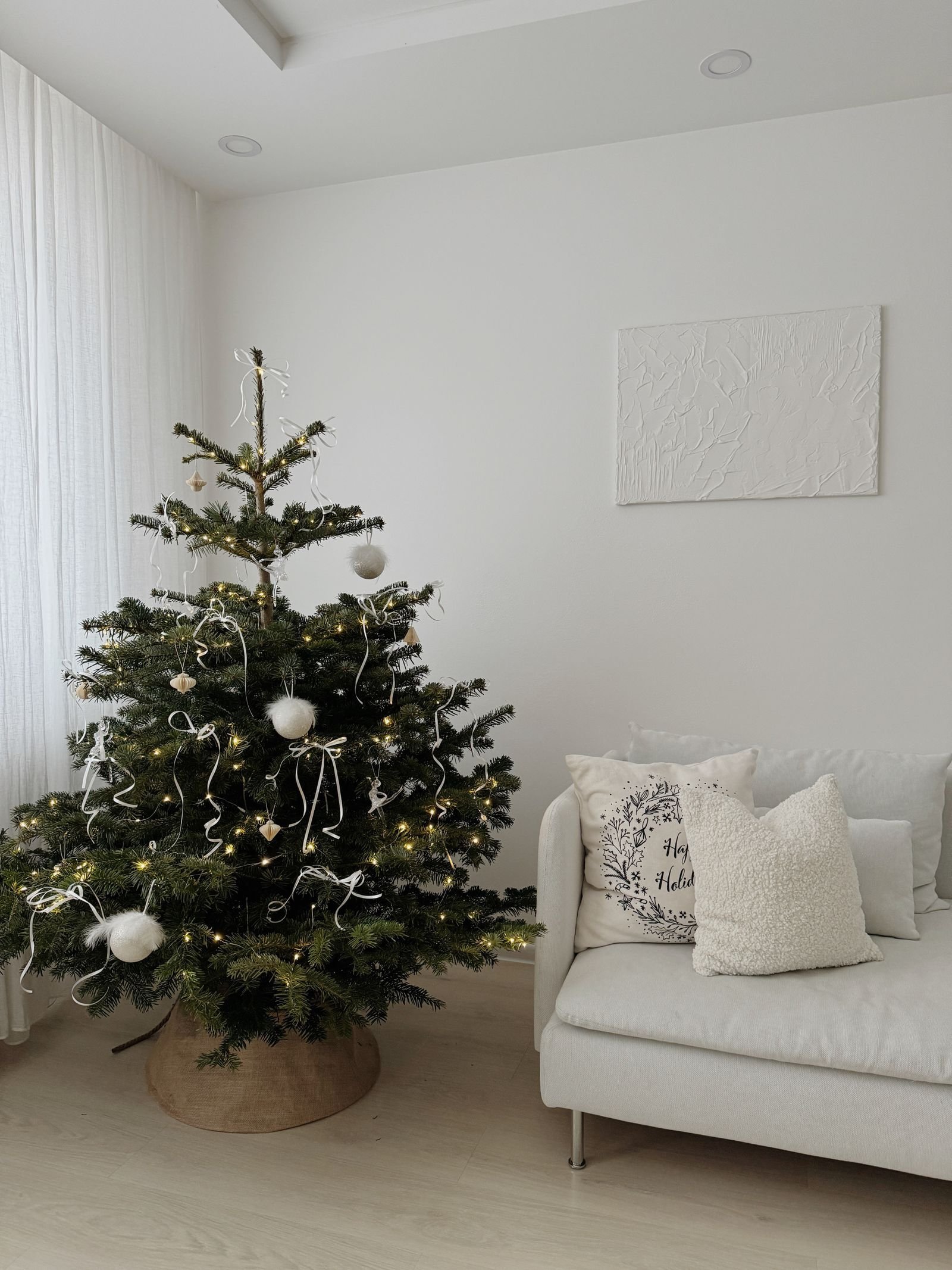A minimalist Christmas tree with white ornaments beside a white sofa in a bright living room