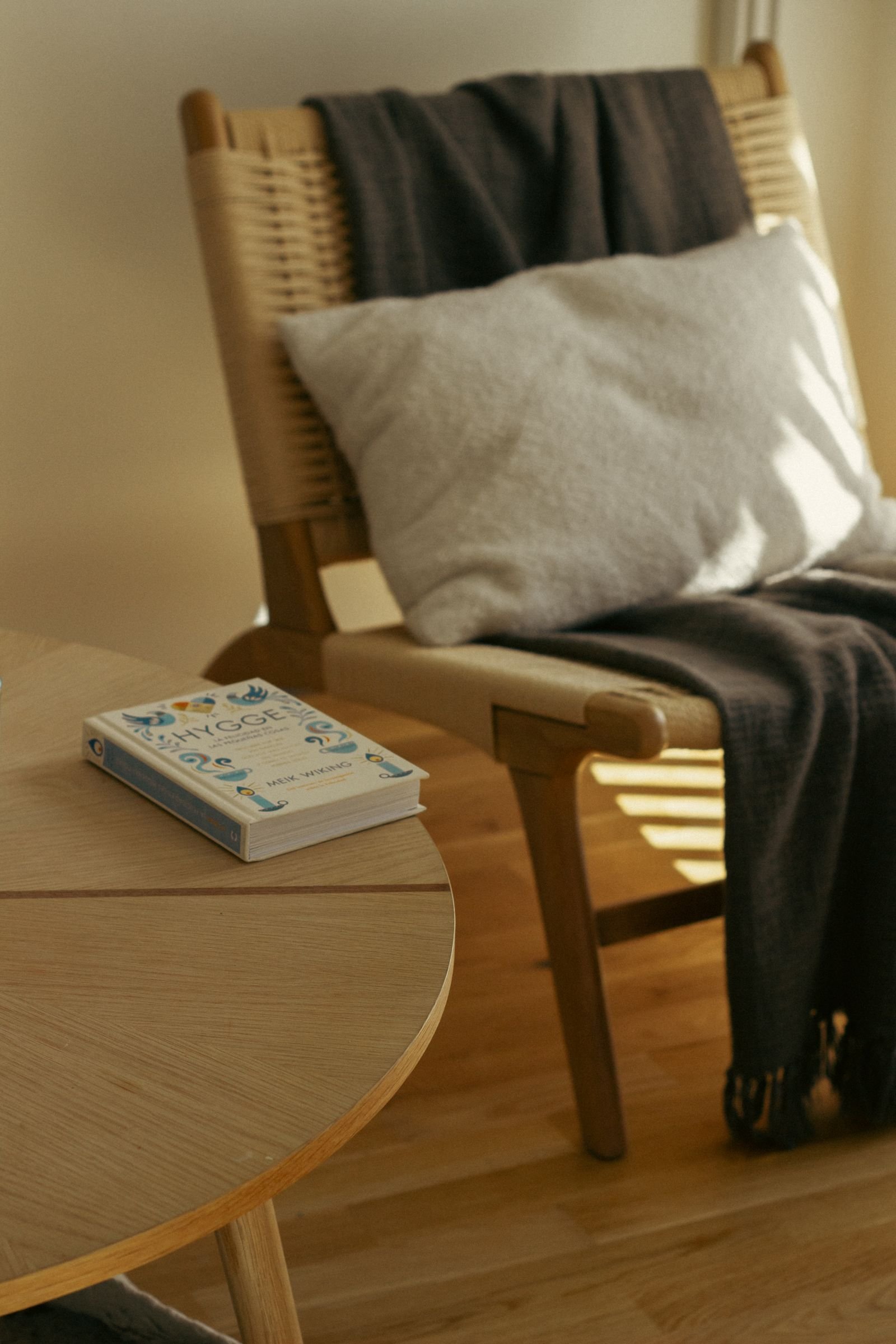 A sunlit reading nook with a wood lounge chair, a gray blanket, a cushion and a book on a round table