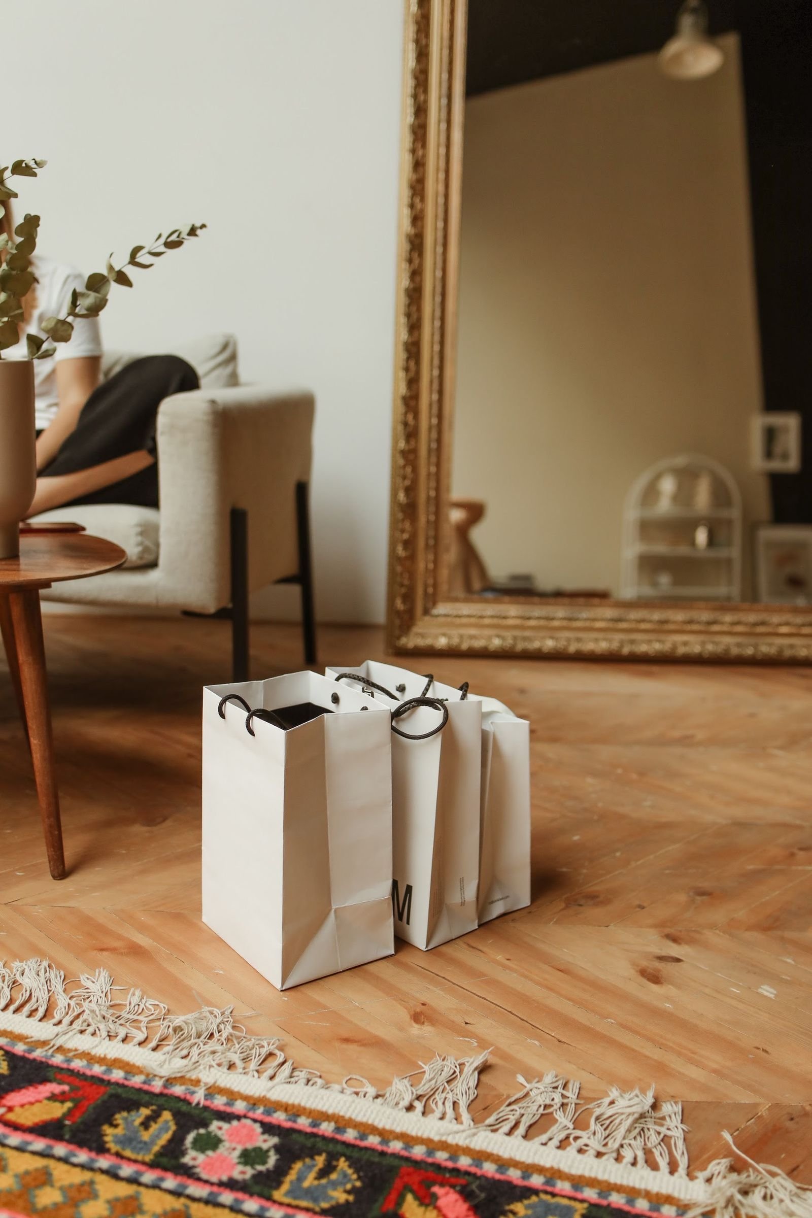 White shopping bags on a wooden floor in a living room with a large ornate mirror and a patterned rug