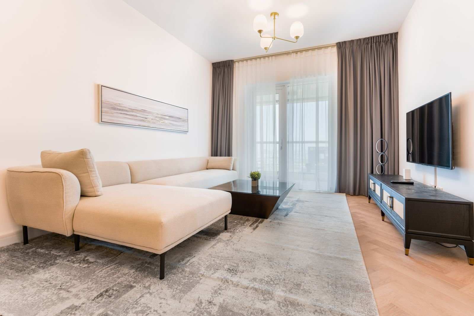A small, light-filled living room with a cream sectional, low TV console and soft gray rug