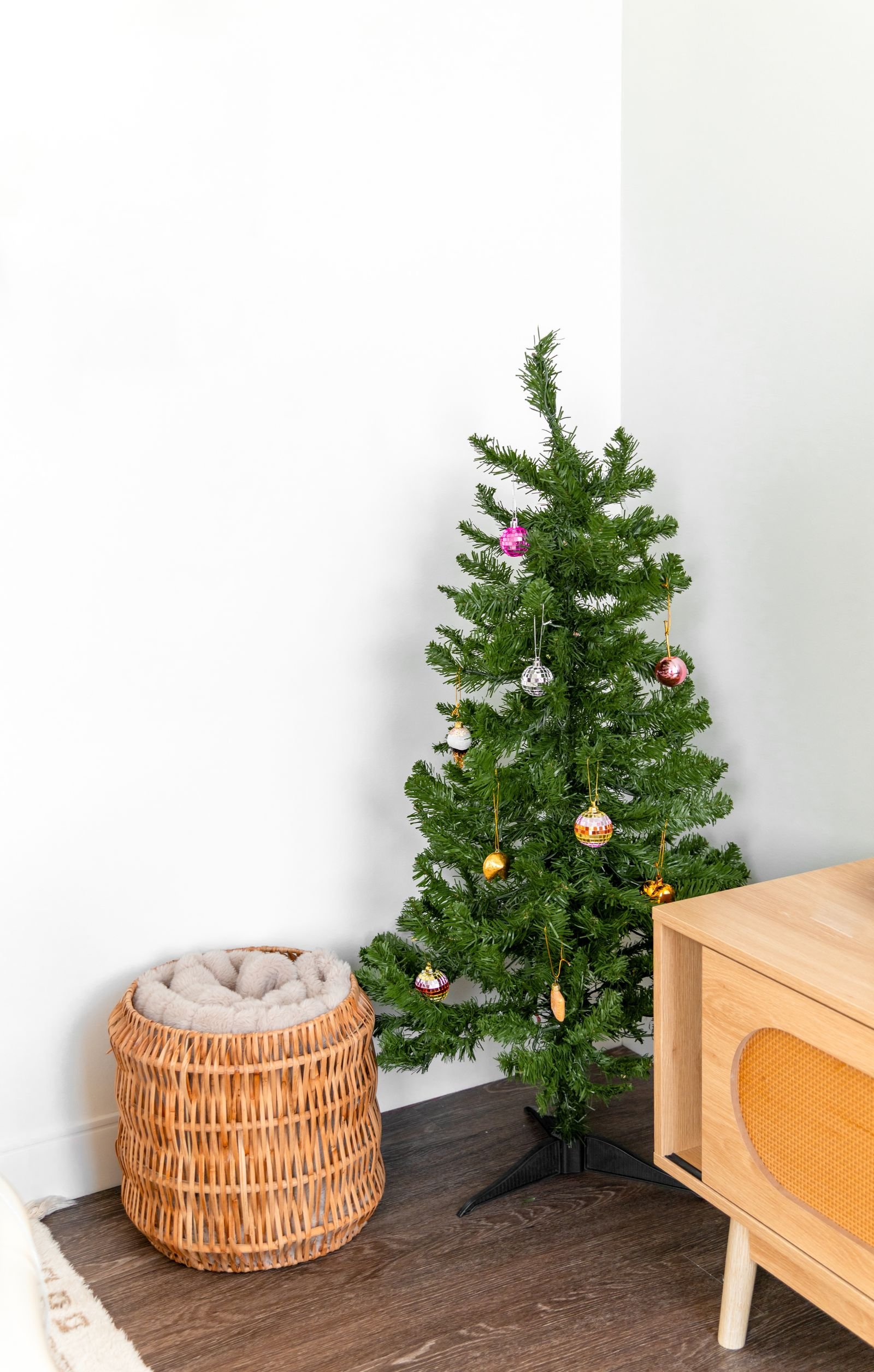 A small decorated Christmas tree beside a woven basket and a wooden cabinet in a bright minimalist room