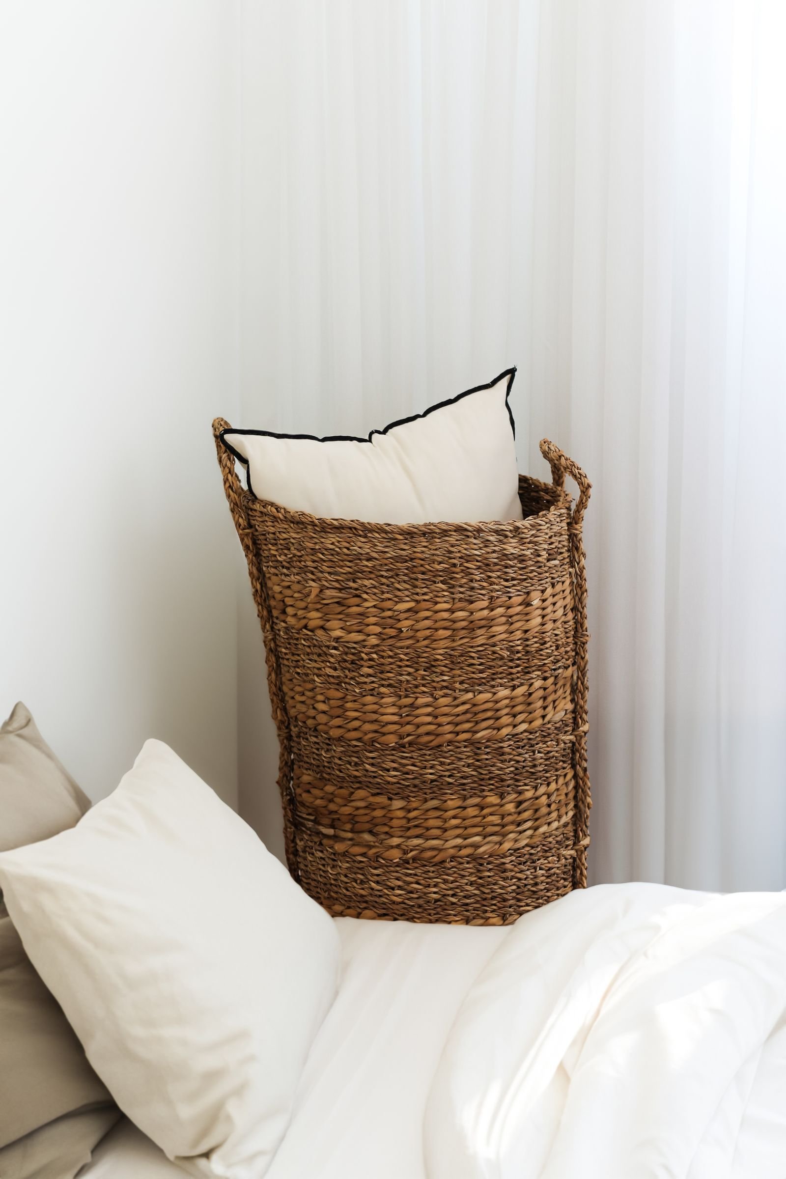 A tall woven wicker basket holding pillows beside a bed with white, neutral bedding