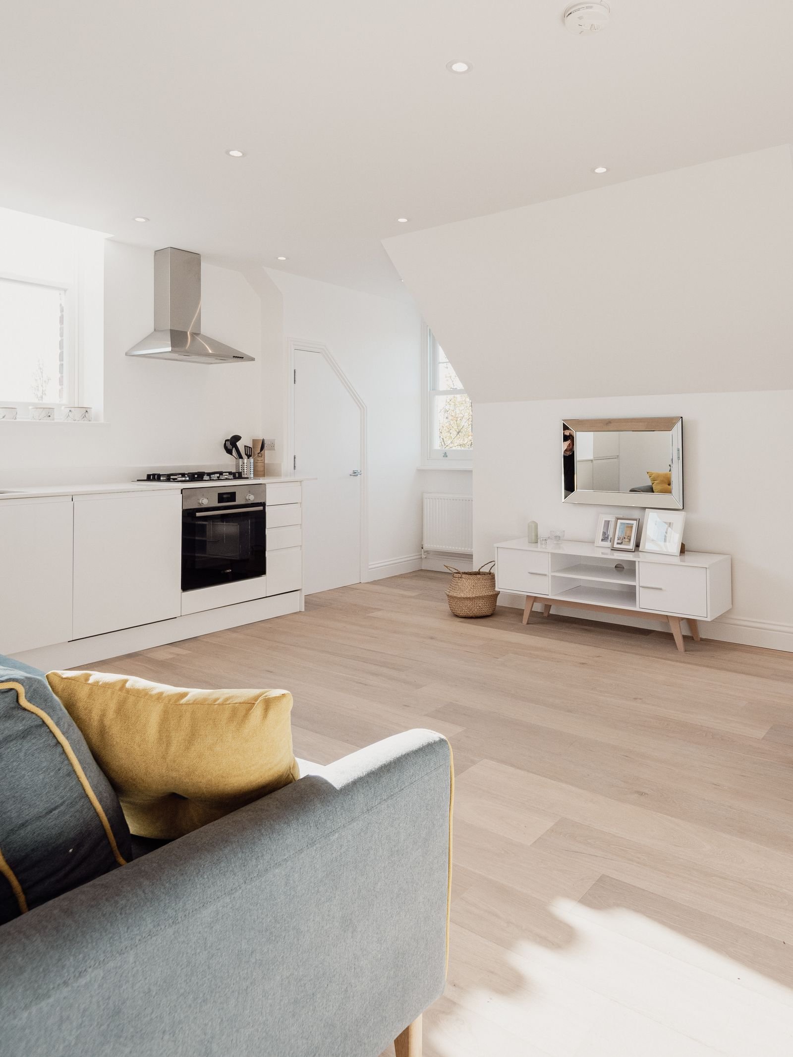 A bright, simple open living space with a gray sofa, yellow pillow, white media console and kitchen behind