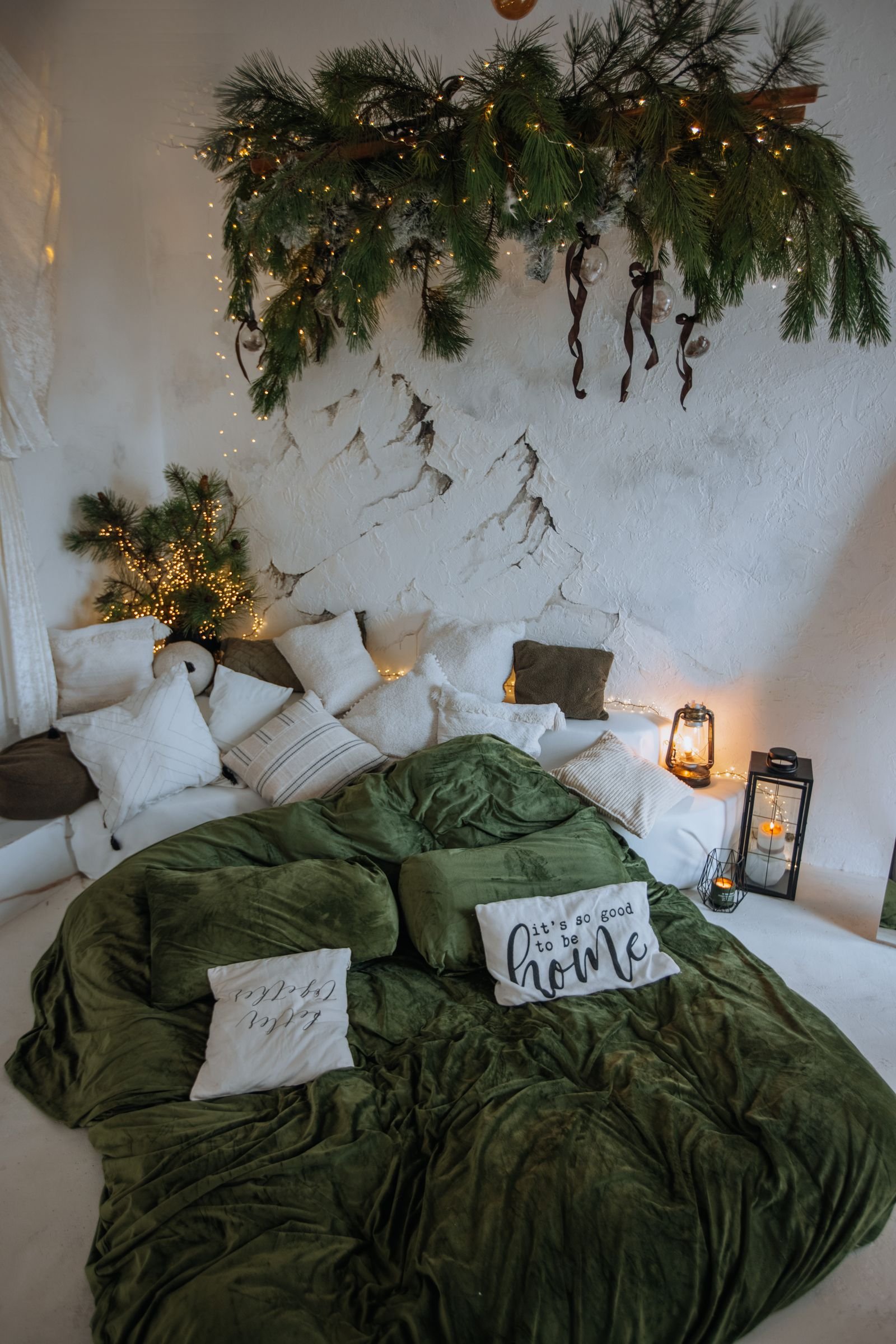 A cozy winter bedroom with a green duvet, white pillows, lanterns and hanging evergreen branches