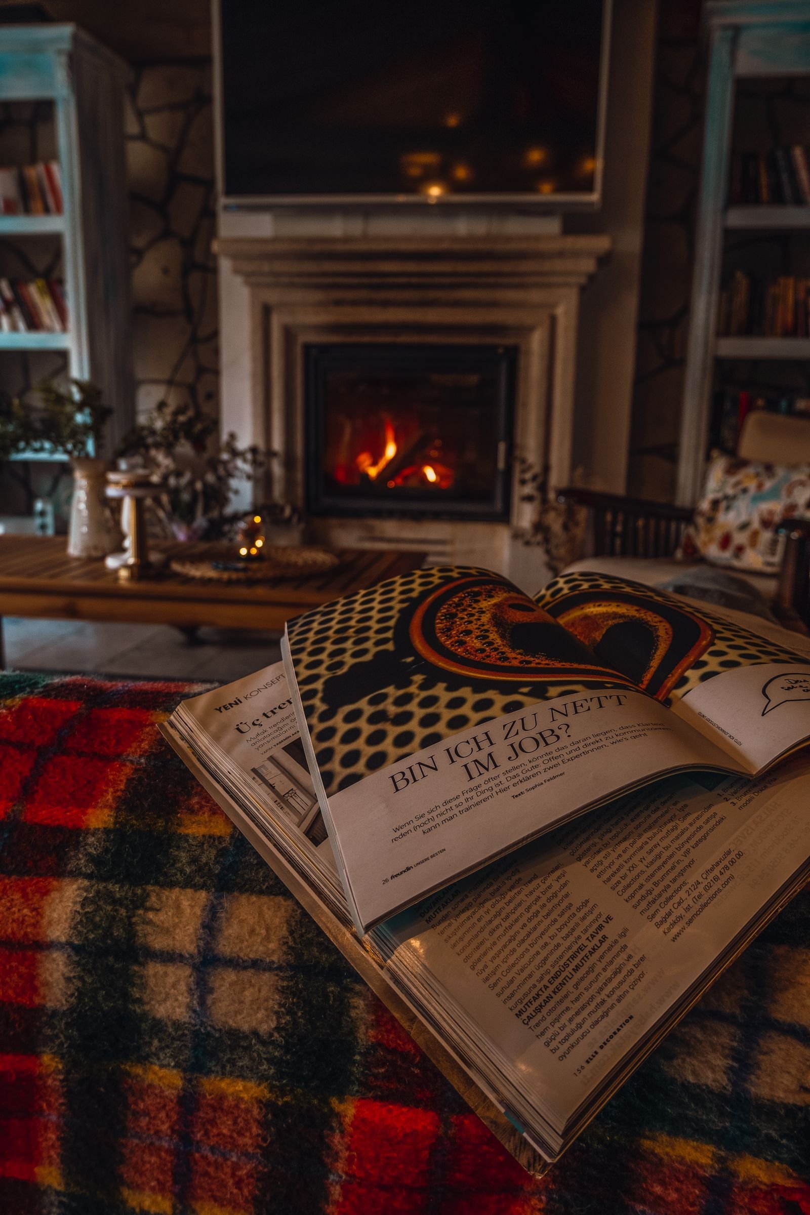A warm living room with a lit fireplace and an open magazine on a plaid blanket